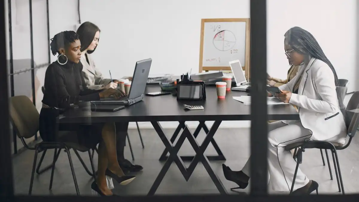 Tiga wanita terlibat dalam kerja tim dan diskusi di sekitar meja di lingkungan kantor modern.