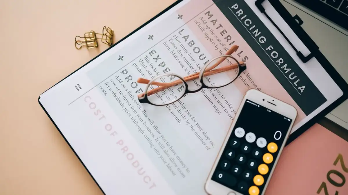 Tampilan dari atas alat bisnis termasuk kalkulator ponsel, dokumen formula harga, dan kacamata di atas meja.