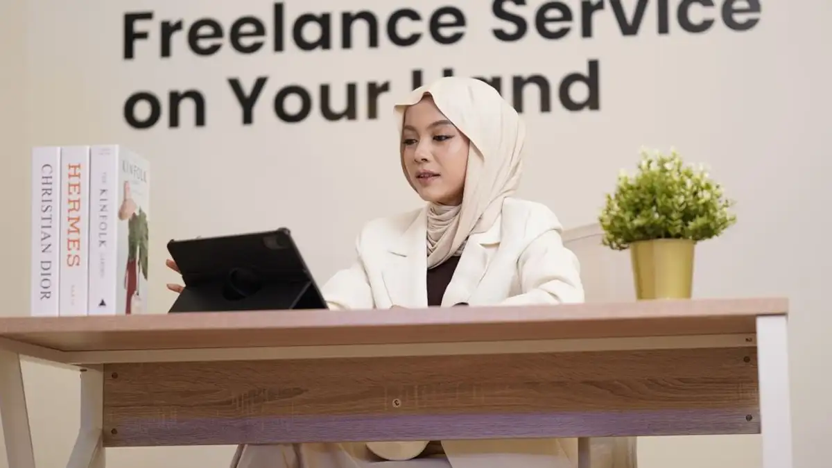 Seorang wanita berhijab sedang bekerja menggunakan tablet di meja yang dilengkapi buku dan tanaman.