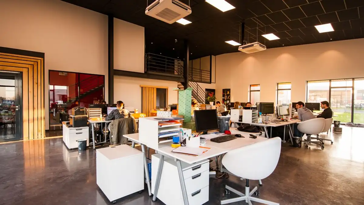 Kantor yang luas dan modern dengan konsep open space, pencahayaan alami, dan karyawan yang bekerja di meja.