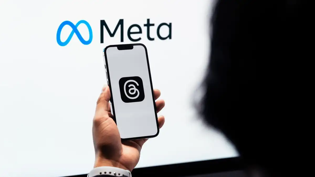 Tangan memegang smartphone dengan logo Threads Meta di layar, branding Meta di latar belakang.