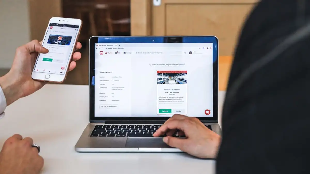 Tangan menggunakan smartphone dan laptop untuk browsing digital di lingkungan kantor, menunjukkan budaya kerja jarak jauh.