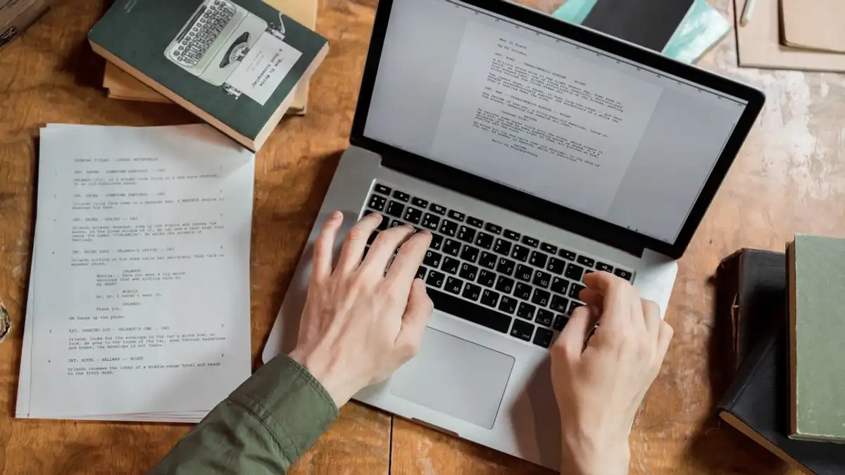 Tampilan sudut tinggi tangan yang mengetik di laptop dikelilingi oleh buku dan kertas.