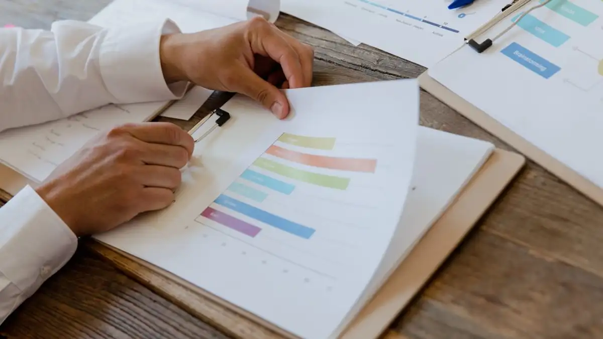Close-up tangan meninjau laporan bisnis dengan grafik dan diagram berwarna di meja kayu.