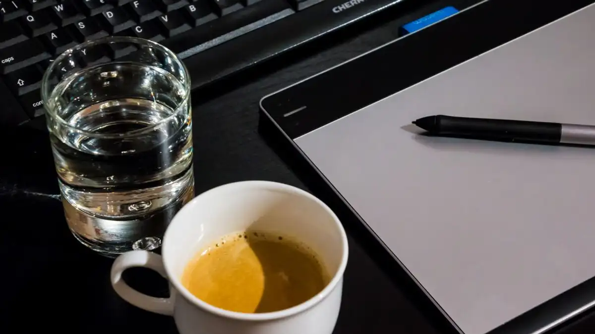 Pengaturan meja kantor dengan cangkir kopi, gelas air, tablet, pena, dan keyboard.