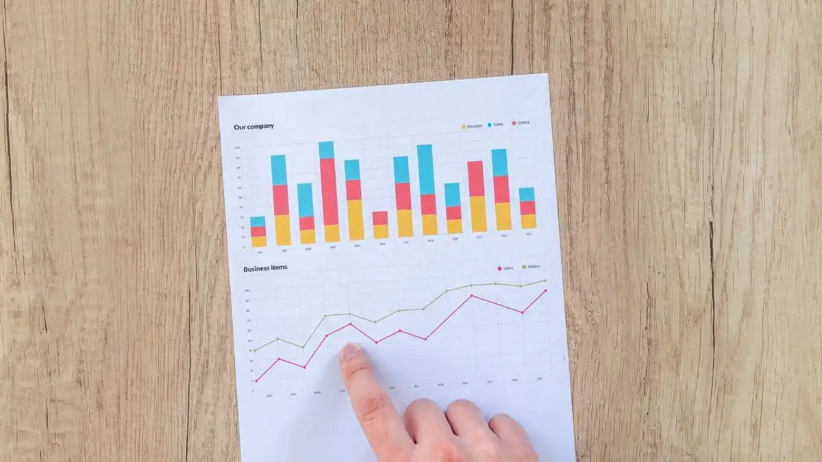 Sebuah tangan menunjukkan grafik dan diagram bisnis berwarna-warni di selembar kertas di atas meja kayu.