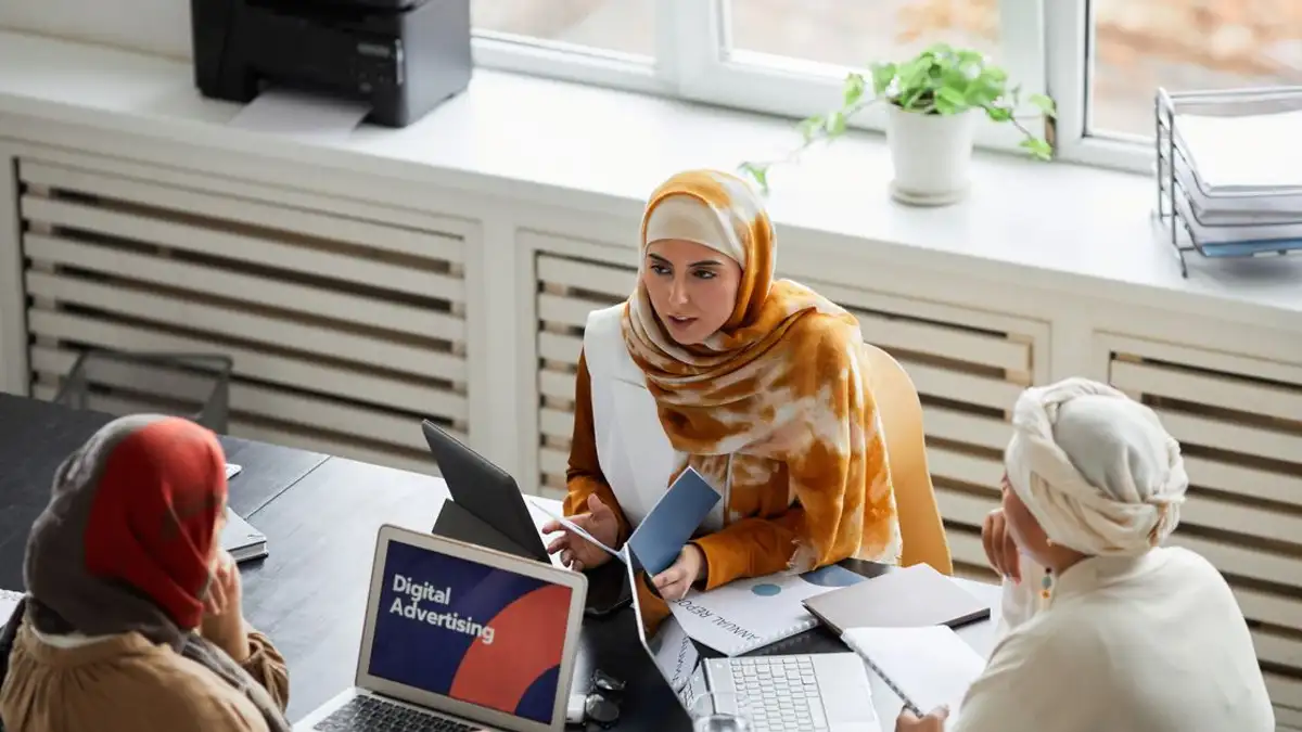 Tiga perempuan pebisnis berpakaian hijab mendiskusikan iklan digital di meja kantor.
