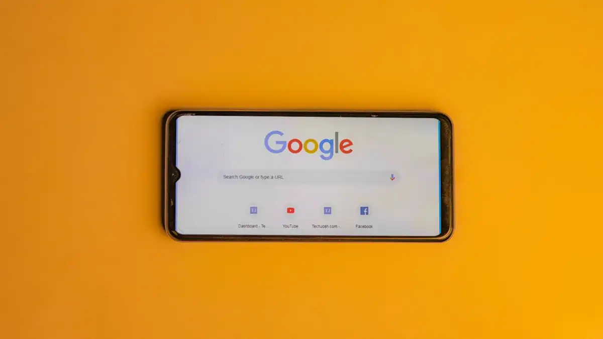 Smartphone menampilkan halaman pencarian Google di latar belakang kuning cerah.