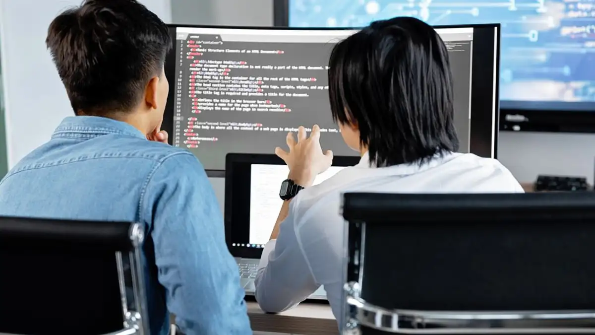 Dua pengembang bekerja sama di kantor, mendiskusikan kode di layar. Lingkungan kerja teknologi.