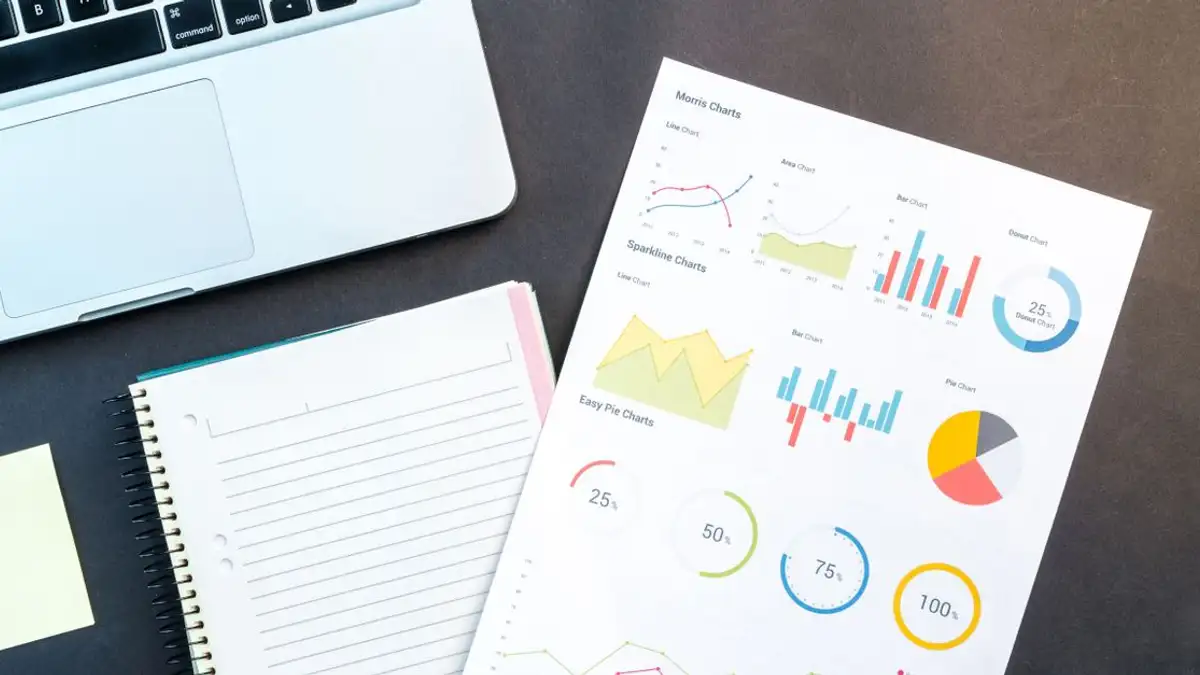 Flatlay laporan bisnis dengan grafik berwarna, buku catatan, dan laptop untuk analisis data di atas meja.