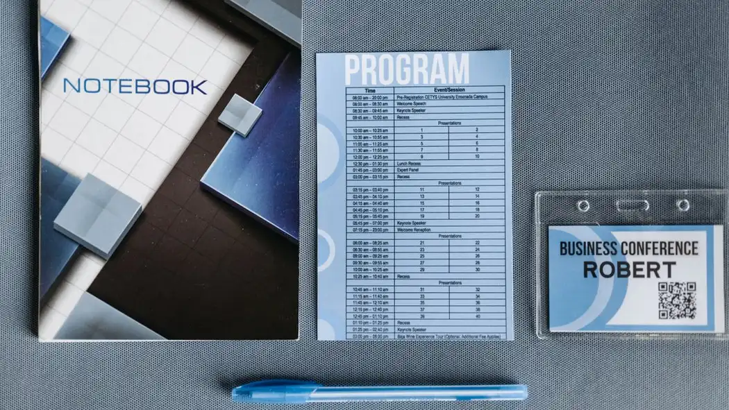 Tampak atas buku catatan, lencana konferensi, pulpen, dan program di permukaan abu-abu.