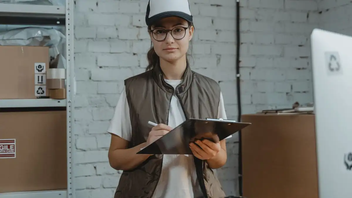 Wanita muda dengan clipboard di gudang, mengelola inventaris dengan presisi.