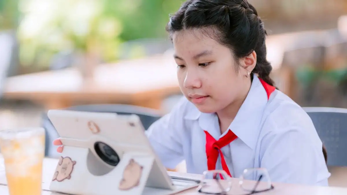 Gadis muda berseragam sekolah belajar di tablet di luar, dengan minuman dan gelas di sampingnya.