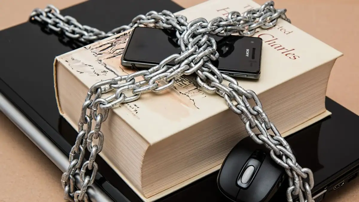 Buku terkunci dengan rantai, ponsel, dan laptop yang melambangkan keamanan digital dan intelektual.