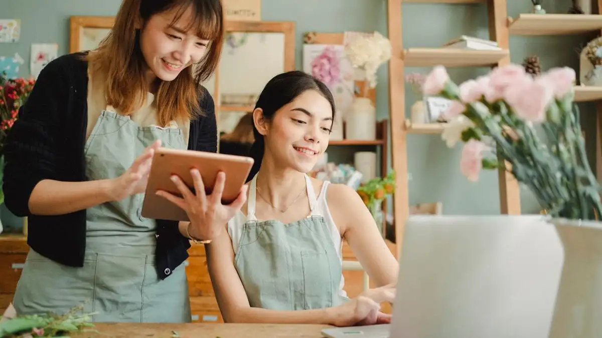 Dua perangkai bunga yang tersenyum bekerja bersama dengan perangkat digital di toko bunga.