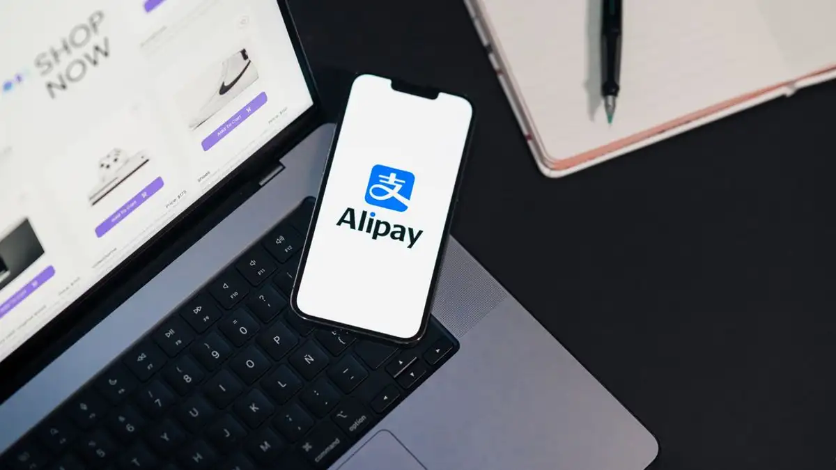 Ponsel pintar menampilkan aplikasi Alipay di laptop terbuka dengan situs belanja daring.