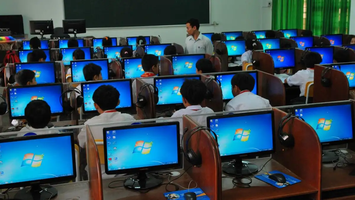 Siswa belajar di laboratorium komputer dengan beberapa monitor dan seorang guru yang mengajar.
